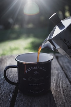 Sunlight filters through trees, creating a soft, glowing effect over a rustic outdoor setting. A metal cup with 'Kathmandu' branding is placed on a weathered wooden surface, and coffee is being poured into it from a metallic coffee maker. The surrounding greenery is out of focus, enhancing the tranquil, natural ambiance.