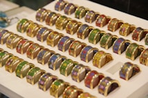 A collection of colorful, intricately designed rings with various patterns, displayed in a grid on a white background. Each ring features unique artwork, including floral and abstract motifs, with gold accents.