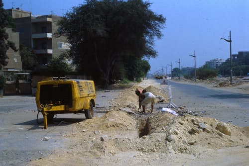 A construction worker wearing shorts and a hat is bent over, working on a trench or excavation site along a road. A large yellow compressor or generator is stationed nearby. The environment appears dusty and there's sparse traffic, with power poles and trees visible along the roadside.