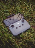 A drone controller with a smartphone attached is resting on a bed of green grass. The controller has two joystick controls and several buttons.