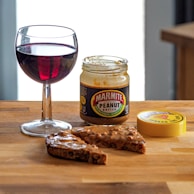 A jar of Marmite Crunchy Peanut Butter sits on a wooden surface next to two slices of bread covered with the peanut butter. A glass of red wine accompanies the setup, creating a contrast between the savory spread and the drink. The jar lid is placed nearby.