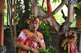 A person dressed in traditional, colorful Balinese attire with detailed headgear and face paint. The background features lush greenery and a tree, with another person similarly dressed standing partially visible.