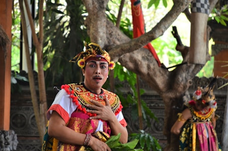 A person dressed in traditional, colorful Balinese attire with detailed headgear and face paint. The background features lush greenery and a tree, with another person similarly dressed standing partially visible.