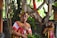 A person dressed in traditional, colorful Balinese attire with detailed headgear and face paint. The background features lush greenery and a tree, with another person similarly dressed standing partially visible.