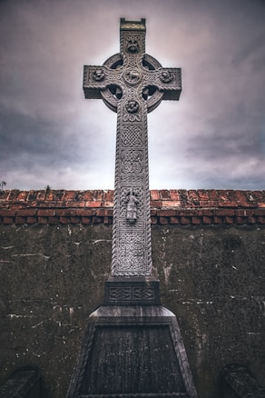 A tall, intricately carved stone Celtic cross stands against a cloudy sky. The cross features detailed patterns and symbols, including circular motifs and figures. It is set against a brick wall and an aged stone base, adding to the historical and solemn atmosphere.