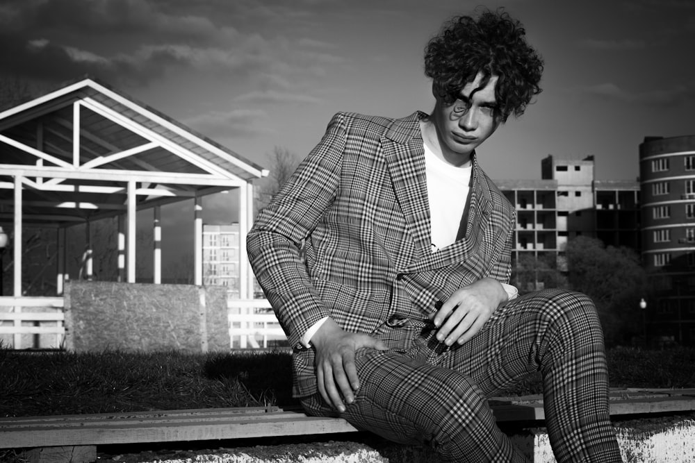 grayscale photo of man in suit jacket and plaid pants sitting on concrete bench