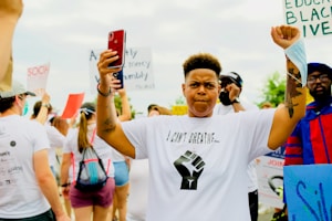 A person with short hair and raised fists stands among a group of protestors, wearing a white T-shirt with the phrase 'I Can't Breathe' and a graphic of a raised fist. Surrounding them are other individuals holding signs with messages supporting social and racial justice. The crowd appears diverse and engaged.