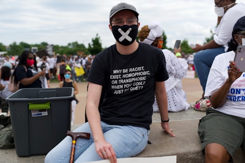 A person sitting on a ledge wearing a black face mask and a black t-shirt with a message against racism, homophobia, and transphobia. The setting appears to be a public demonstration or protest, with people in the background wearing masks and holding signs. The atmosphere suggests a socially conscious gathering.