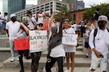 A diverse group of people are gathered at a protest holding signs advocating for racial justice. Some signs read 'My Blackness isn't a crime' and 'Black Lives Matter'. The expression of the crowd is energetic and determined, with people raising their fists in solidarity.