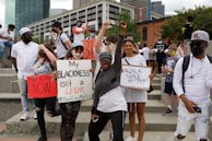 A diverse group of people are gathered at a protest holding signs advocating for racial justice. Some signs read 'My Blackness isn't a crime' and 'Black Lives Matter'. The expression of the crowd is energetic and determined, with people raising their fists in solidarity.