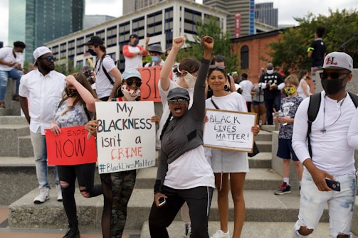 A diverse group of people are gathered at a protest holding signs advocating for racial justice. Some signs read 'My Blackness isn't a crime' and 'Black Lives Matter'. The expression of the crowd is energetic and determined, with people raising their fists in solidarity.