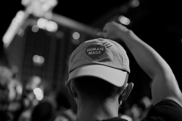 A grayscale photograph of a person from behind wearing a cap with the words 'HUMAN MADE' on it. The individual has their arm raised, possibly in a fist. The background is blurred with various lights and an indistinct cityscape visible.