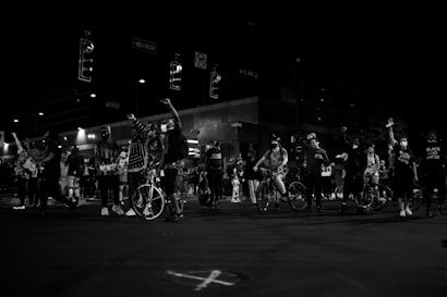 A group of people, some with bicycles, gather on a street at night. Many individuals have their fists raised and are wearing masks. Signs and traffic lights are visible above, indicating an urban environment. Some individuals wear clothing with messages advocating for racial justice.