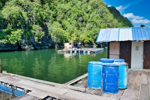 A rustic wooden platform over a body of green water, with a small building that has a blue and white metal roof. Several large blue barrels are positioned on the wooden planks. In the background, lush green foliage covers steep rocky cliffs, and a small floating structure is tethered to the platform.