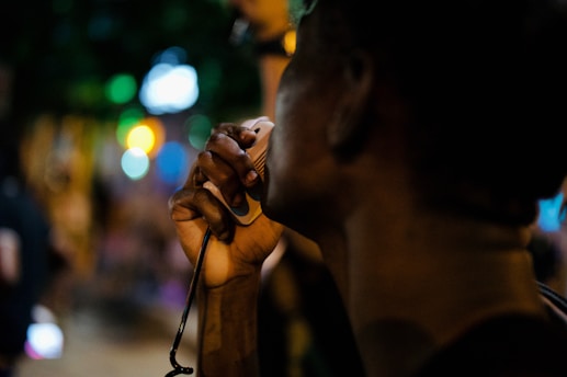 A person is speaking into a handheld microphone in an outdoor setting at night. The background is blurred with bokeh lights in various colors, suggesting a lively or crowded atmosphere.