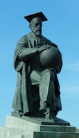 A bronze statue depicts a man wearing a long academic robe and a mortarboard hat, sitting with one leg crossed over the other. The figure holds a globe-like object in his hands, and the base of the statue is a stone platform. The background features a clear blue sky.