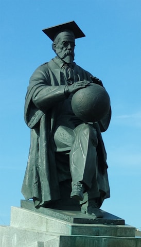 A bronze statue depicts a man wearing a long academic robe and a mortarboard hat, sitting with one leg crossed over the other. The figure holds a globe-like object in his hands, and the base of the statue is a stone platform. The background features a clear blue sky.