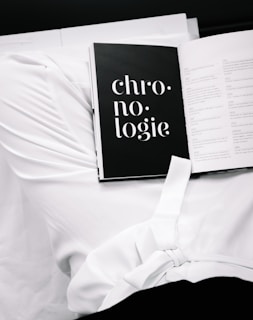 An open book displaying a page with the word 'chronologie' in bold white typography on a black background. The book rests on a rumpled white fabric, suggesting a casual or intimate setting.