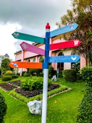 A colorful signpost stands in a well-maintained garden with various directional signs featuring phrases about love. Each sign is painted in bright colors and points in different directions. The background includes a building with large arched windows and a manicured lawn with neatly trimmed bushes and flowers. The sky is overcast with gray clouds.