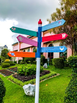 A colorful signpost stands in a well-maintained garden with various directional signs featuring phrases about love. Each sign is painted in bright colors and points in different directions. The background includes a building with large arched windows and a manicured lawn with neatly trimmed bushes and flowers. The sky is overcast with gray clouds.