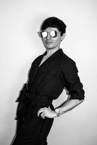 A person poses confidently wearing reflective sunglasses and a dark outfit. Their posture is stylish, with one hand on the hip and the other relaxed. The background is plain and minimalistic.