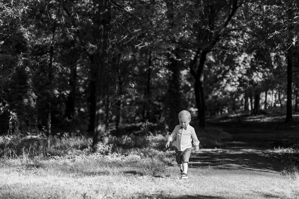 grayscale photo of child walking on pathway surrounded by trees