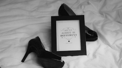 A black and white photograph features a pair of glossy high-heeled shoes and a framed quote displayed on a textured fabric surface. The quote reads 'You must always be different' attributed to Coco Chanel. The arrangement suggests a theme of elegance and individuality.