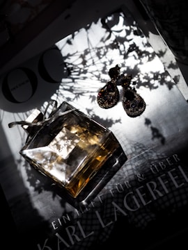 A stylish composition featuring a Chanel perfume bottle and a pair of elegant earrings with intricate designs, placed on a surface that appears to be a magazine featuring the name 'Karl Lagerfeld'. The scene is illuminated with dramatic shadows from nearby foliage.