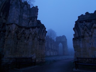An atmospheric scene depicting ancient stone ruins enveloped in a deep blue mist. The arches and detailed stonework suggest a Gothic architectural style. Two wooden benches are positioned symmetrically within the image. Tall leafless trees are visible in the background, adding to the eerie and tranquil atmosphere.
