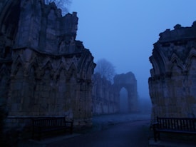 An atmospheric scene depicting ancient stone ruins enveloped in a deep blue mist. The arches and detailed stonework suggest a Gothic architectural style. Two wooden benches are positioned symmetrically within the image. Tall leafless trees are visible in the background, adding to the eerie and tranquil atmosphere.
