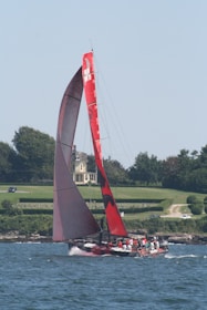 A large red sailboat glides through the water with several people on board, wearing life vests. The boat features a prominent red sail with markings. A picturesque landscape with a grand house, neatly trimmed hedges, and trees serves as the backdrop.
