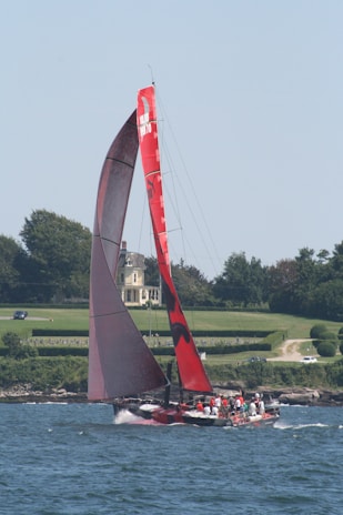 A large red sailboat glides through the water with several people on board, wearing life vests. The boat features a prominent red sail with markings. A picturesque landscape with a grand house, neatly trimmed hedges, and trees serves as the backdrop.