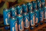 Multiple blue cans featuring images and text in an organized display, possibly representing a religious or spiritual theme. The design on the cans includes a depiction of a figure resembling religious icons, and the text is in a foreign language, suggesting a cultural context.