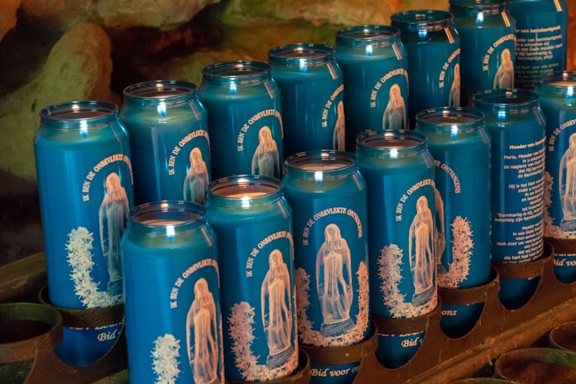 Multiple blue cans featuring images and text in an organized display, possibly representing a religious or spiritual theme. The design on the cans includes a depiction of a figure resembling religious icons, and the text is in a foreign language, suggesting a cultural context.