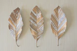 Three dried leaves with gold and white geometric patterns are displayed against a light wooden background. The designs are intricate, featuring a mix of lines, circles, and triangles, creating a tribal or bohemian aesthetic.