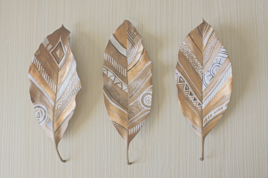 Three dried leaves with gold and white geometric patterns are displayed against a light wooden background. The designs are intricate, featuring a mix of lines, circles, and triangles, creating a tribal or bohemian aesthetic.