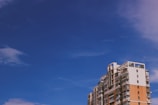 A modern, multi-story apartment building with a mix of white and orange brick facade stands against a clear blue sky. The architecture features balconies and large windows.
