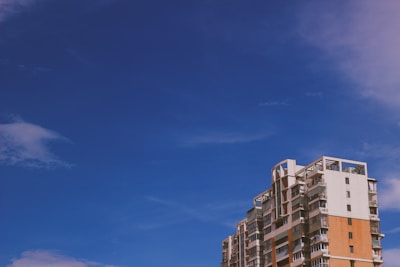 A modern, multi-story apartment building with a mix of white and orange brick facade stands against a clear blue sky. The architecture features balconies and large windows.
