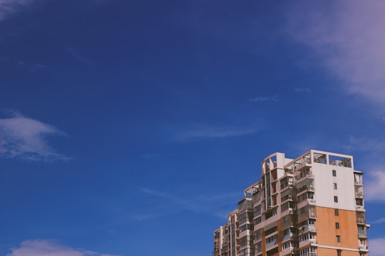 A modern, multi-story apartment building with a mix of white and orange brick facade stands against a clear blue sky. The architecture features balconies and large windows.