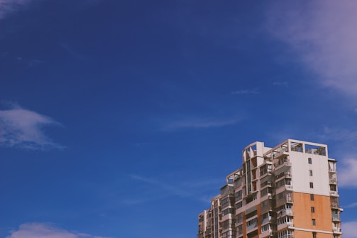 A modern, multi-story apartment building with a mix of white and orange brick facade stands against a clear blue sky. The architecture features balconies and large windows.