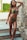 A person in a colorful bikini poses confidently beside a textured stone wall. The backdrop includes steps made of light blue tiles and a lush green garden with a white wall.