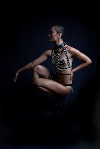 A person is posed against a dark backdrop, wearing a black harness and minimal clothing. The lighting highlights their muscular build and the intricate design of the harness. Their head is shaved, and they're squatting, creating an emphasis on balance and strength.