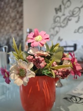 A bright orange vase holds a bouquet of artificial flowers, including pink and white blossoms with green leaves and small pink berry-like clusters. The background features a patterned wall and faint, decorative designs with some blurred elements.