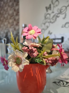A bright orange vase holds a bouquet of artificial flowers, including pink and white blossoms with green leaves and small pink berry-like clusters. The background features a patterned wall and faint, decorative designs with some blurred elements.
