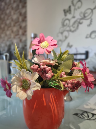 A bright orange vase holds a bouquet of artificial flowers, including pink and white blossoms with green leaves and small pink berry-like clusters. The background features a patterned wall and faint, decorative designs with some blurred elements.