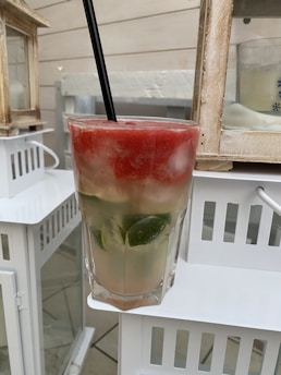 A tall glass filled with a layered beverage featuring red, white, and green colors, topped with ice and a black straw. The drink appears to contain slices of lime and is placed on a white surface with a wooden frame and lanterns in the background.