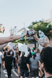 A group of people are participating in a protest or demonstration, holding up signs. Hands reaching upwards are holding water bottles and snack bags. The scene is outdoors, and there is a crowd in the background, showing solidarity and unity.