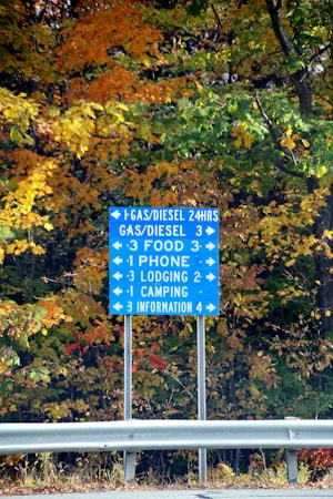 A blue highway information sign is prominently displayed against a backdrop of vibrant autumn foliage. The sign provides directions to various amenities including gas stations, food, phone services, lodging, camping, and information centers. The overall scene is framed by the changing colors of the leaves, which range from green to golden yellow and orange, typical of a fall landscape.
