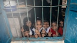 A group of young children smiling through a window with metal bars. The children appear cheerful and lively, looking directly at the viewer. The blue window frame contrasts with the children's colorful clothing.