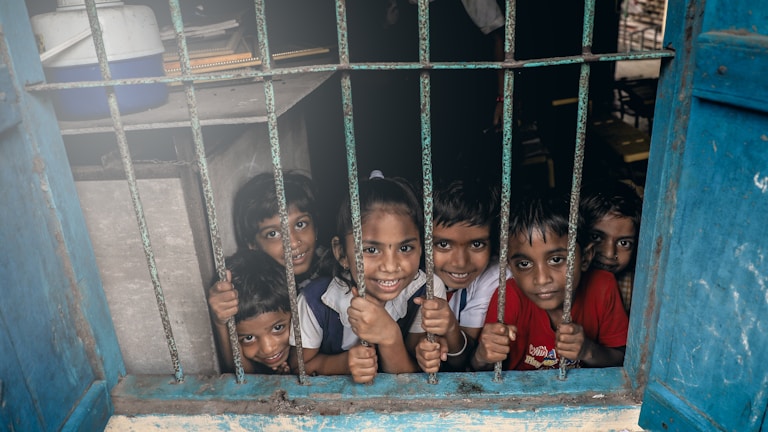 A group of young children smiling through a window with metal bars. The children appear cheerful and lively, looking directly at the viewer. The blue window frame contrasts with the children's colorful clothing.